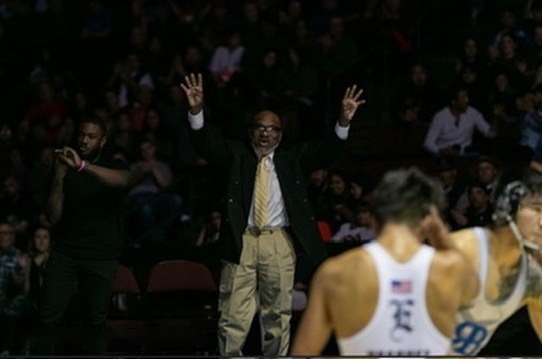 A coach in a suit with both hands raised at the edge of the mat during a match