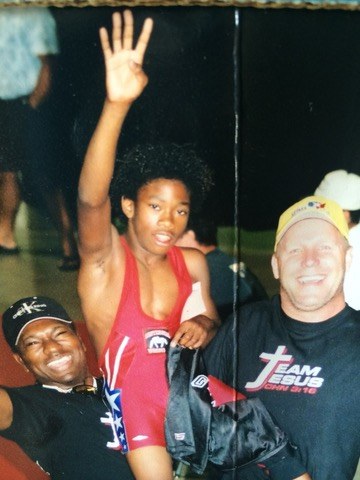 A young wrestler in a red singlet raising one arm between two smiling coaches